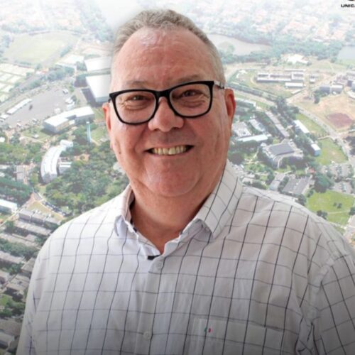 paulo cesar montagner de camisa branca quadriculada e óculos sorrindo com um fundo de uma foto aerea da unicamp
