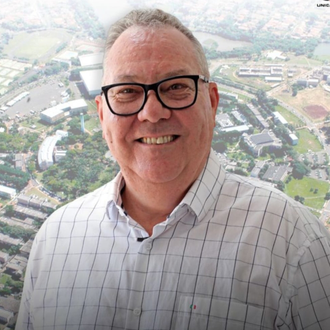 paulo cesar montagner de camisa branca quadriculada e óculos sorrindo com um fundo de uma foto aerea da unicamp